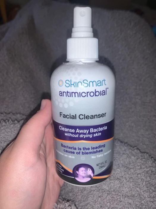 A hand holding a bottle of SkinSmart Antimicrobial Facial Cleanser, promoting its ability to clear bacteria, reduce blemishes, and keep skin moisturized.