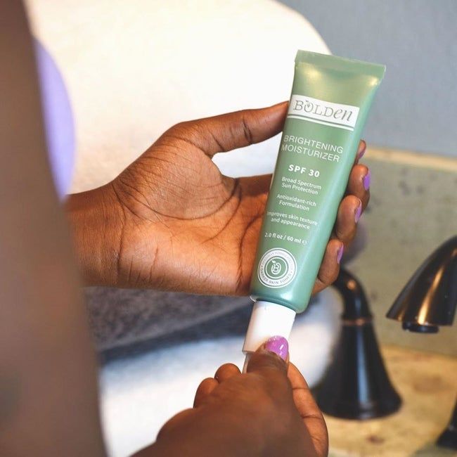 a model holding and opening a green and white tube of Bolden's SPF 30 Brightening Moisturizer