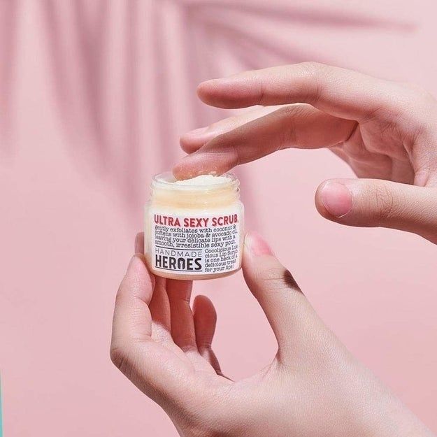 A finger dips into a small transparent jar to gather a beige-colored lip exfoliant