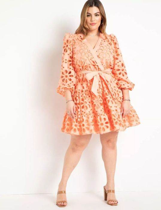 An orange-peach eyelet mini dress is worn by a model paired with tan block heels.