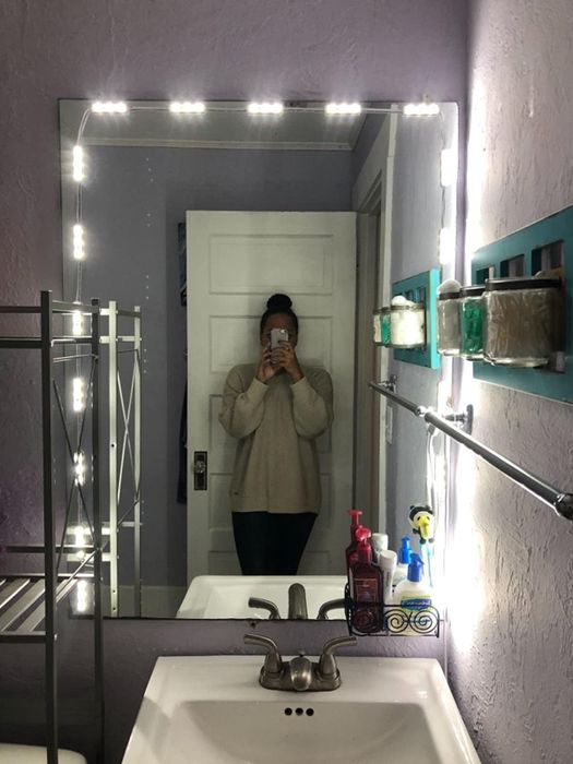 A reviewer takes a selfie in front of a bathroom vanity mirror, adorned with LED lights.