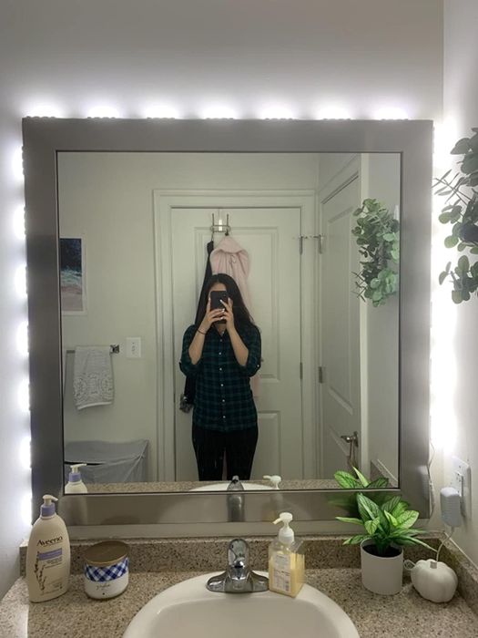 The reviewer stands in front of a square vanity mirror, which is surrounded by the same lights around the edges.