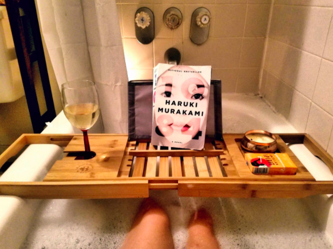 The reviewer’s light wood bath caddy, featuring a glass of white wine, a book, and a candle, is placed over their bathtub.