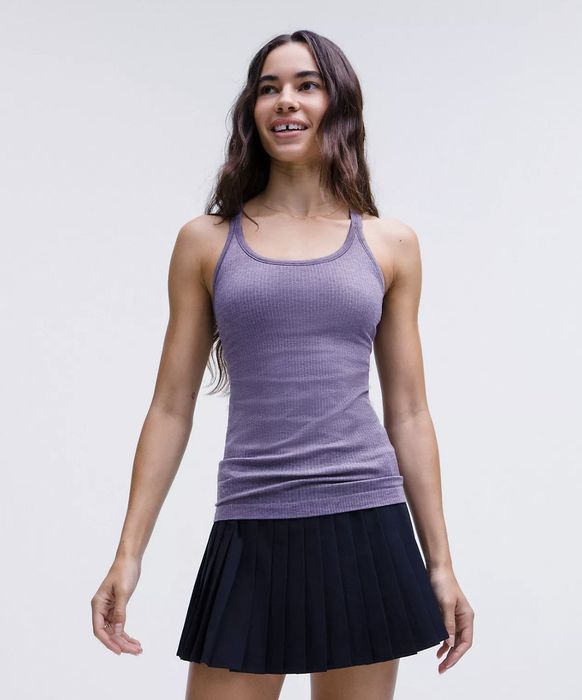 A model wearing a purple fitted tank top and a pleated skirt, smiling while standing against a simple backdrop.