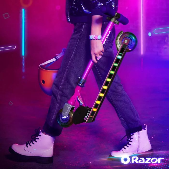 A person holds a colorful Razor scooter, wearing jeans and white boots, with a helmet in the other hand