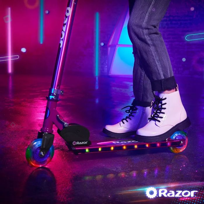 A person rides a Razor scooter with glowing wheels in a modern indoor setting. White boots and jeans are visible, with the Razor logo on the floor