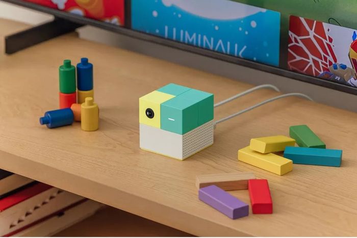 A set of colorful geometric blocks and a small cube-shaped electronic device sits on a wooden shelf, surrounded by bright and playful children's toys.