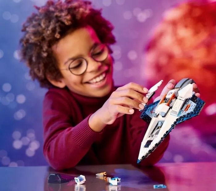 A smiling child builds a toy spaceship, with extra pieces scattered on the table. Caption: