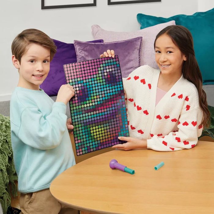 Two children at a table, smiling and holding a colorful art pegboard. A crafting tool is nearby