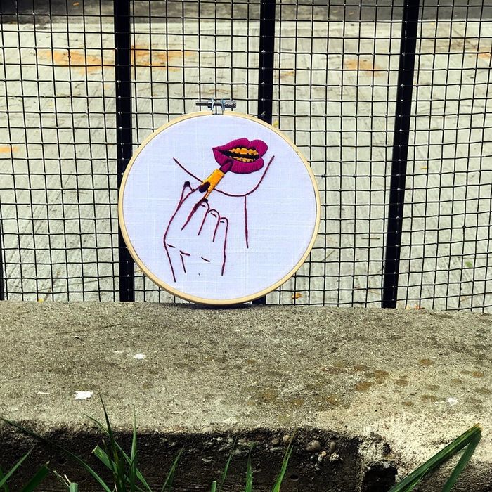 An embroidery hoop featuring a finished design of a person's chin, with red lipstick and a visible gold grill on their teeth