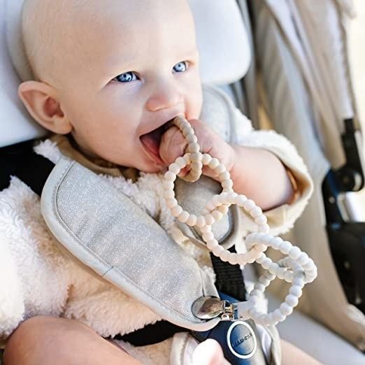 A baby model happily chewing on the teething toy, which is clipped securely to their carseat strap.