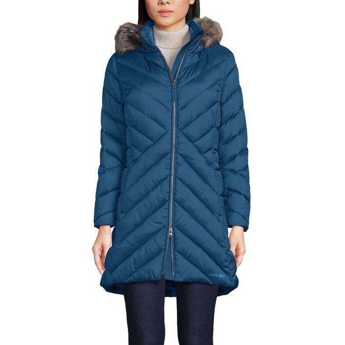A person is wearing a long, dark blue zip-up insulated coat with angled quilting and a faux fur-trimmed hood.