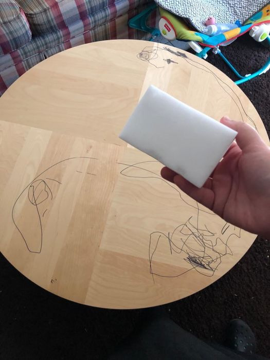 User holding a cleaning eraser sponge in front of a wooden table marked with permanent marker stains
