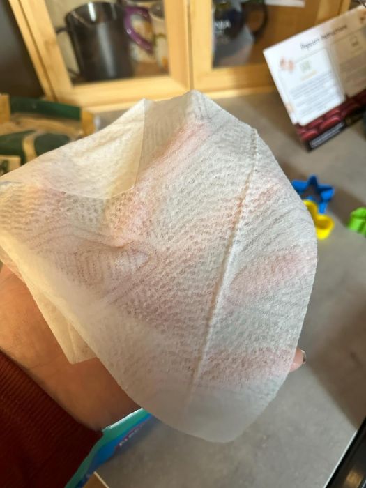 A hand holding a disposable cleaning wipe, with children's toys and a product information leaflet visible on a counter in the background