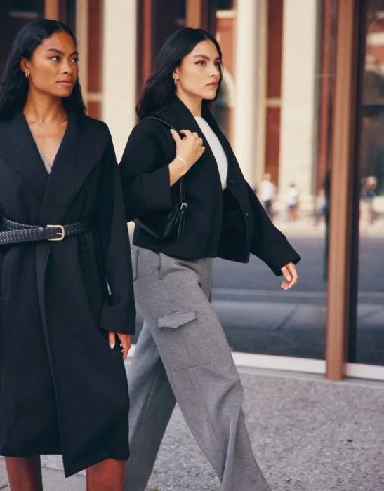 A stylish individual strolls down the avenue, donning gray trousers paired with a sleek black jacket.