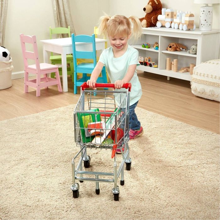 A young girl pushing a metal shopping cart filled with pretend food items.