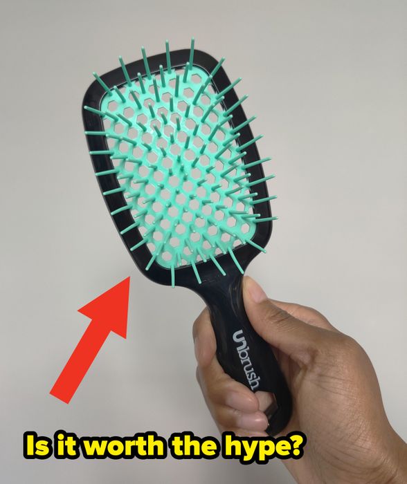 A person holding a black hairbrush with aqua bristles in front of a neutral backdrop