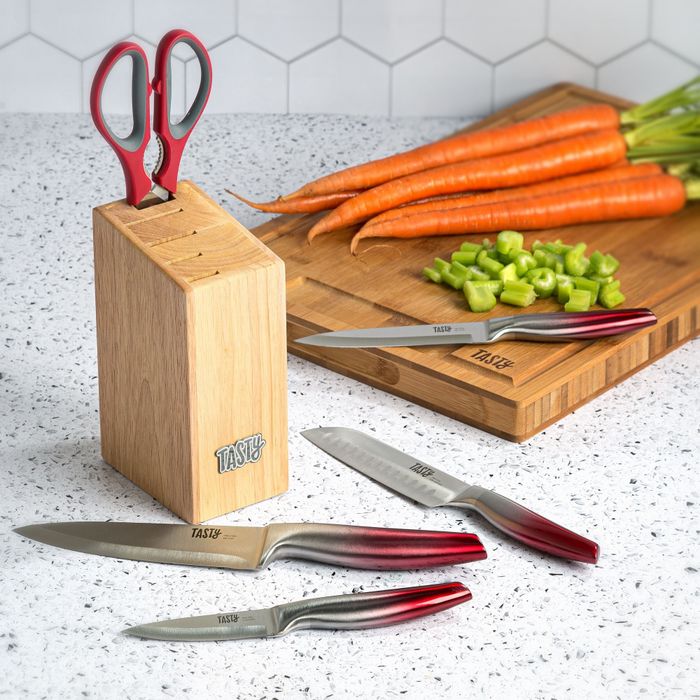 Wooden knife block with red scissors inside, four sharp knives laid out, cutting board with chopped carrots and celery in the background