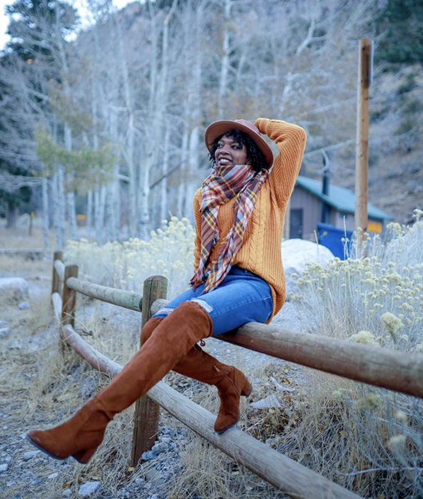 A customer photo of them wearing the sweater in ginger, paired with boots and a hat, sitting comfortably on a wooden fence.