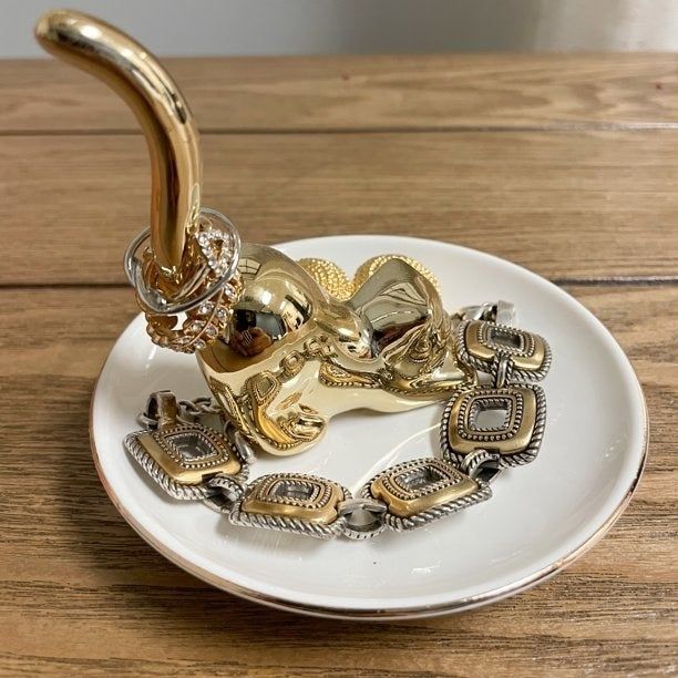 A white dish filled with gold jewelry, featuring a gold cat in the center that holds the rings