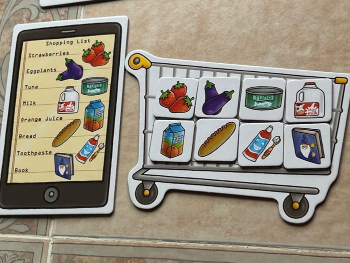 A shopping list displayed on a tablet alongside a magnetic toy shopping cart. Items featured include strawberries, eggplants, tuna, milk, orange juice, bread, toothpaste, and a book.