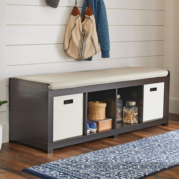 An espresso wood colored storage bench with a beige cushion, flanked by two white cubes on either side, with open cubbies in the middle filled with various items