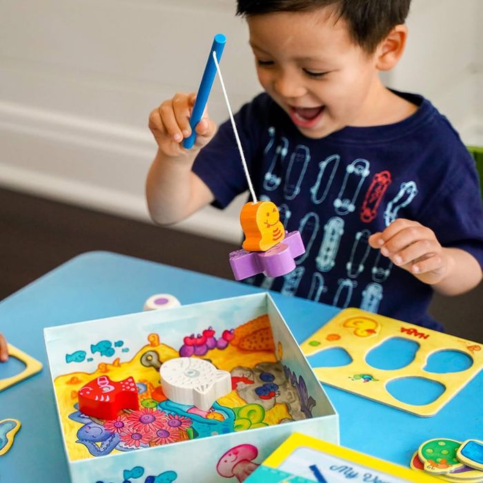A child happily plays with a fishing game, using a rod to catch wooden fish from a colorful box set