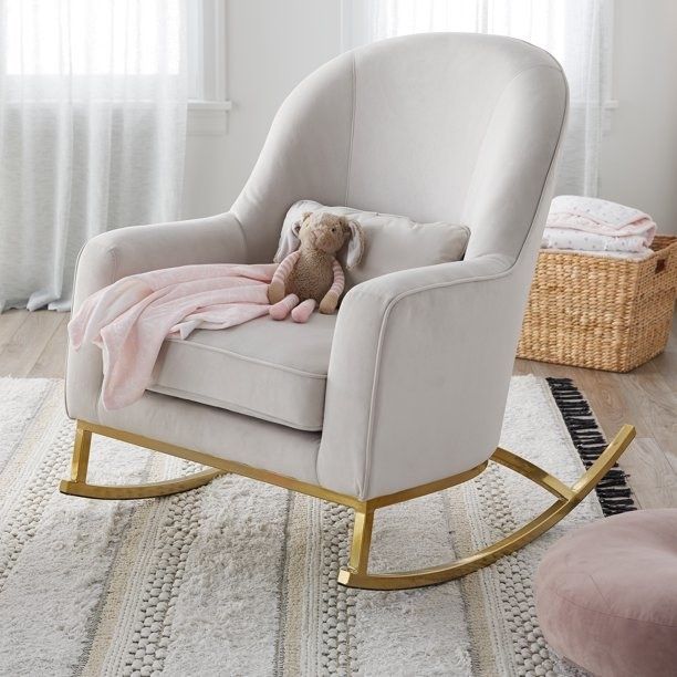 A beige rocking chair with a gold rocker base, a pink blanket, and a teddy bear resting on it.
