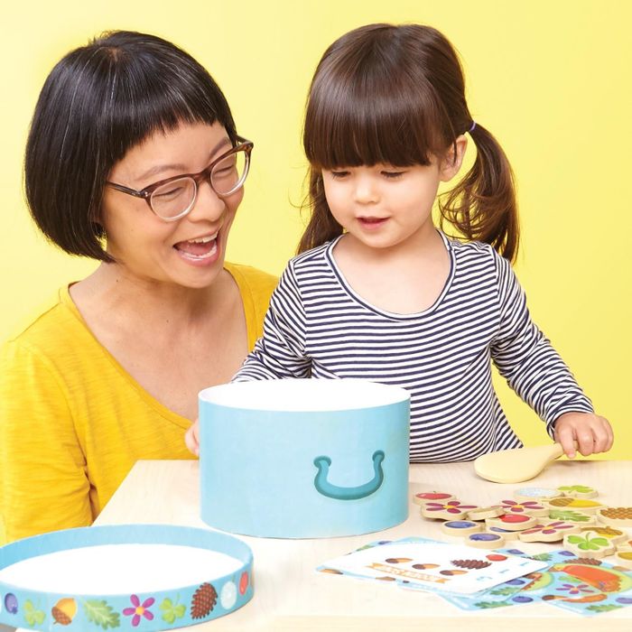 An adult and child are deeply engrossed in playing with a Montessori-style toy on a table, sorting shapes into a container. Their expressions radiate joy and focus.