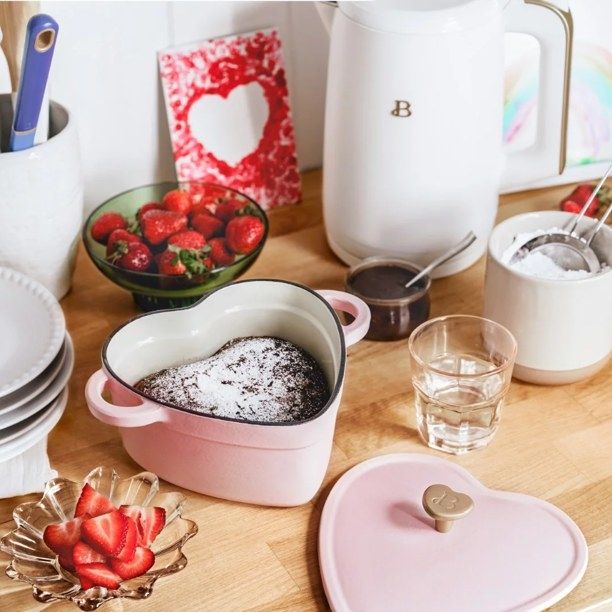A pink heart-shaped Dutch oven containing a chocolate cake, surrounded by a heart-shaped lid, strawberries, and glassware on a table.