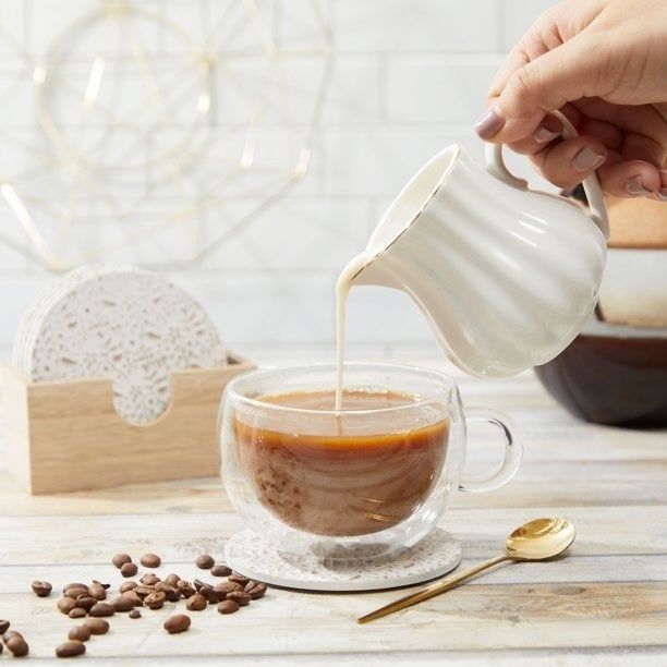 A model pouring milk into a glass mug of coffee, which rests on a white coaster.