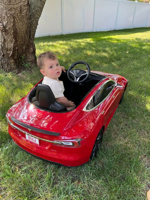 A toddler sitting in a mini Tesla Model S ride-on car, glancing over their shoulder