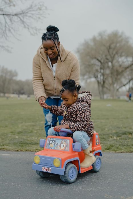An adult helping a child in a toy car, both casually dressed and outdoors