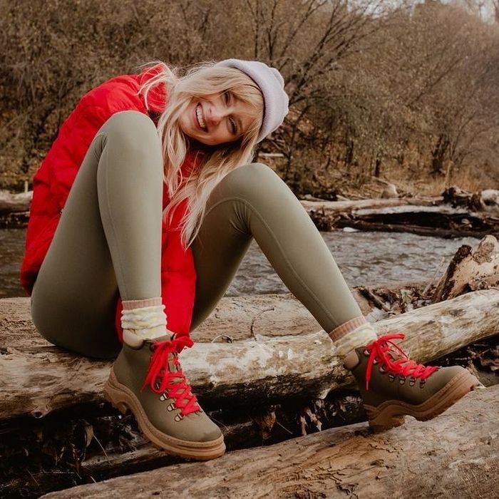 A model styled in a puffer jacket, green leggings, and the khaki green boots with red laces, sitting on a log