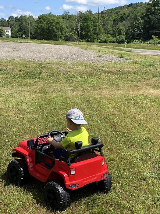 A child in a toy car, sporting a bucket hat and casual clothes, riding outdoors on the grass