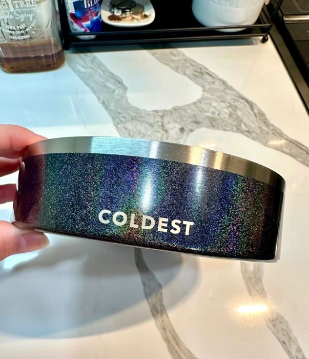 A person is holding a shiny metallic bowl emblazoned with the word "COLDEST" on it. The bowl reflects a brilliant holographic finish.