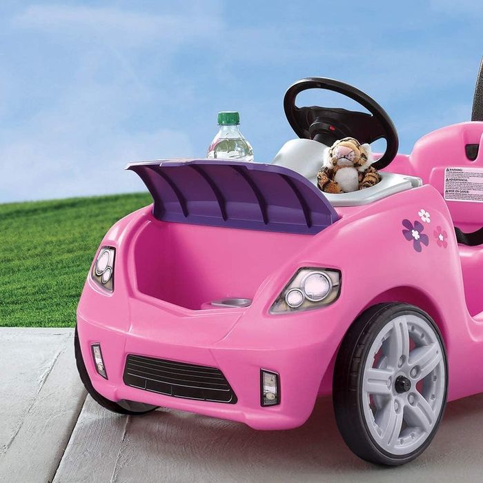 A pink toy car parked on the grass, with a water bottle and stuffed toy resting on the pavement