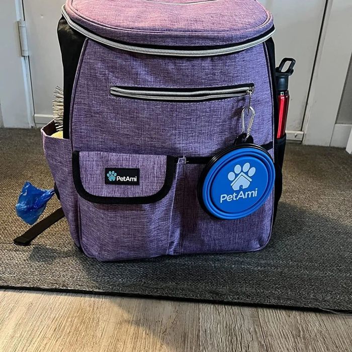A purple PetAmi backpack, featuring front pockets, a water bottle holder, and a circular PetAmi logo tag. The photo is taken indoors.