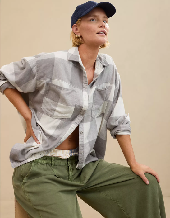 A person in a relaxed outfit, wearing a plaid shirt, green pants, and a cap, posing against a neutral backdrop