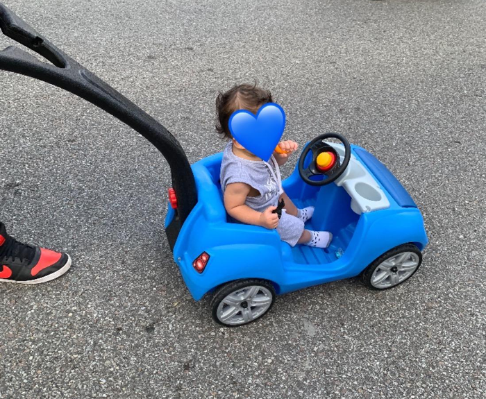 A child riding in a blue toy car stroller on a sidewalk, with an adult walking alongside