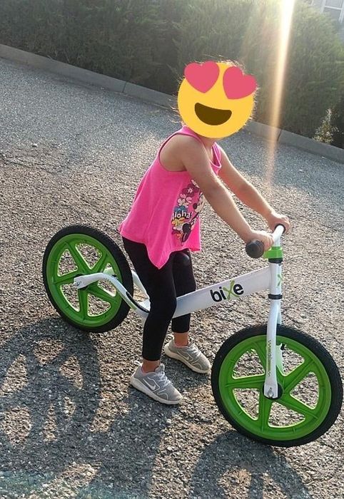 A child riding a balance bike, covering their face with a heart emoji, wearing a tank top and shorts