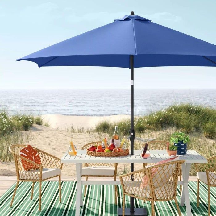 A green striped outdoor rug, paired with a white table, rattan chairs, and a blue umbrella, set on a beach with a view of the water