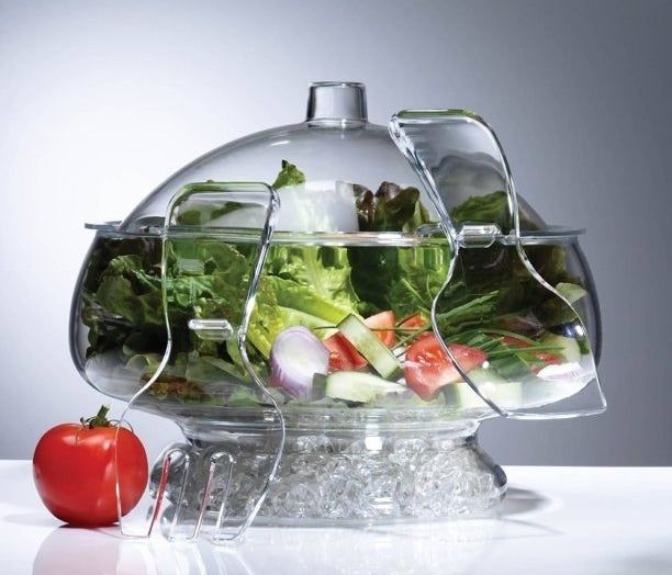 The glass salad bowl, with ice at the bottom and greens on top, covered by a matching domed lid and salad tongs hanging on the side.