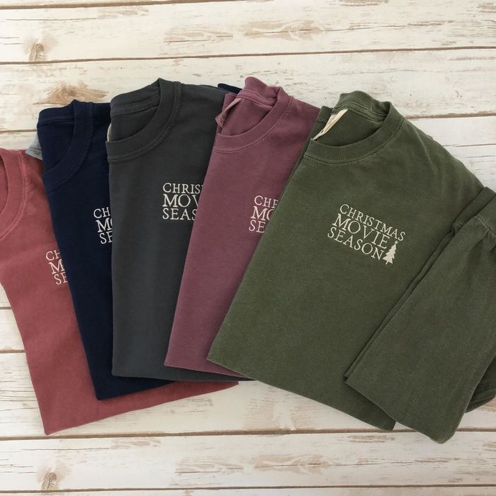 five cozy sweatshirts in shades of red, blue, grey, maroon, and green, featuring the phrase "christmas movie season" alongside a festive tree design
