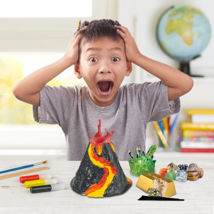A young child eagerly examines a model volcano kit on a table, surrounded by DIY materials and geological samples.