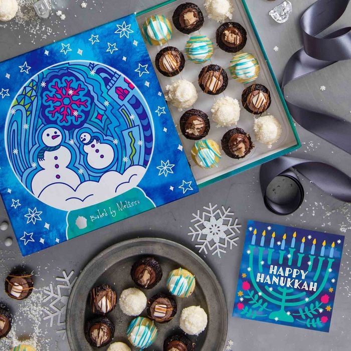 A box adorned with blue and pink snowman motifs containing vibrant mini cupcakes, accompanied by a Hanukkah-themed card displaying a menorah and the greeting 'Happy Hanukkah'.