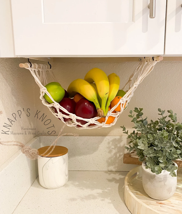 A fruit hammock holding bananas and apples, hanging under a cabinet with a plant on a round wooden board on the kitchen counter nearby