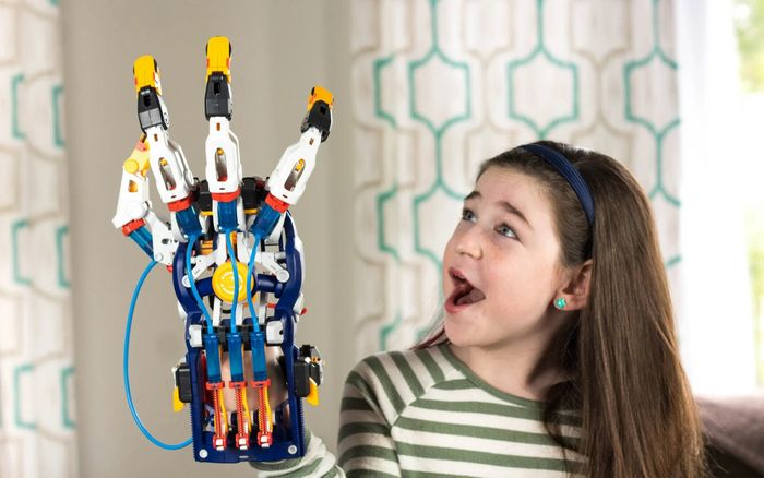 A young child, dressed in a striped shirt, gazes in awe at a robotic hand toy she is holding up
