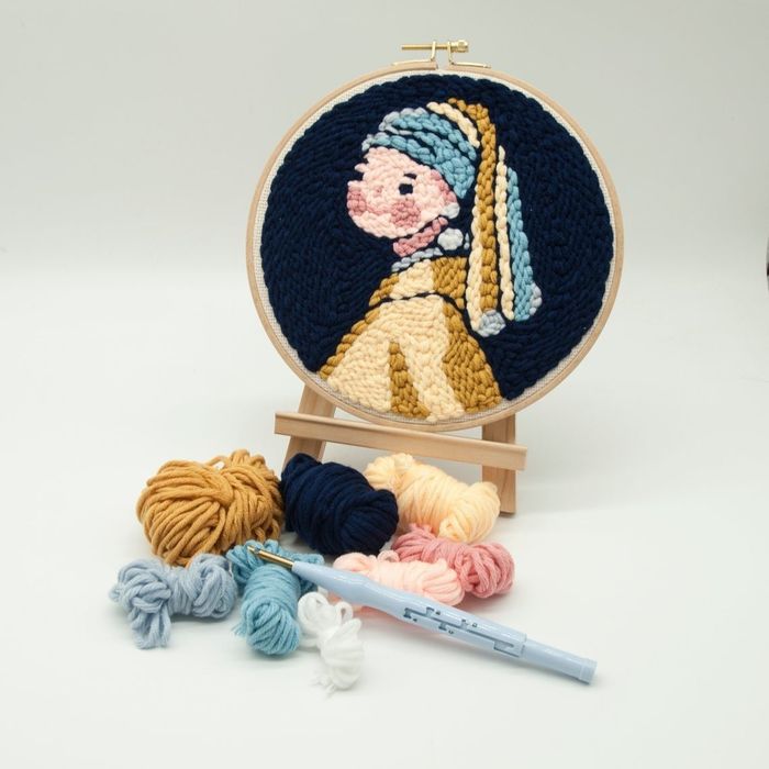 an embroidery hoop showcasing a chunky, cartoon-style depiction of the girl with a pearl earring, accompanied by yarn and a punch needle