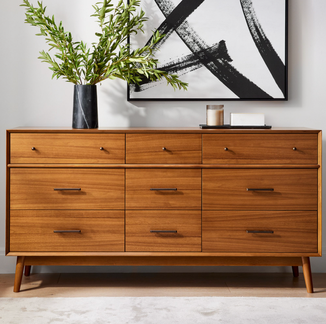 A wooden dresser with six spacious drawers, sleek handles, and angled legs, placed in a room with a plant and wall art.